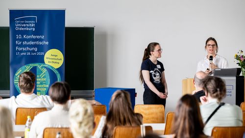 Zwei junge Frauen stehen hinter und neben einem Rednerpult, links befindet sich ein Banner mit dem Logo der Konferenz. Man sieht von hinten einige Personen auf Stühlen sitzen, die den beiden zuhören. 