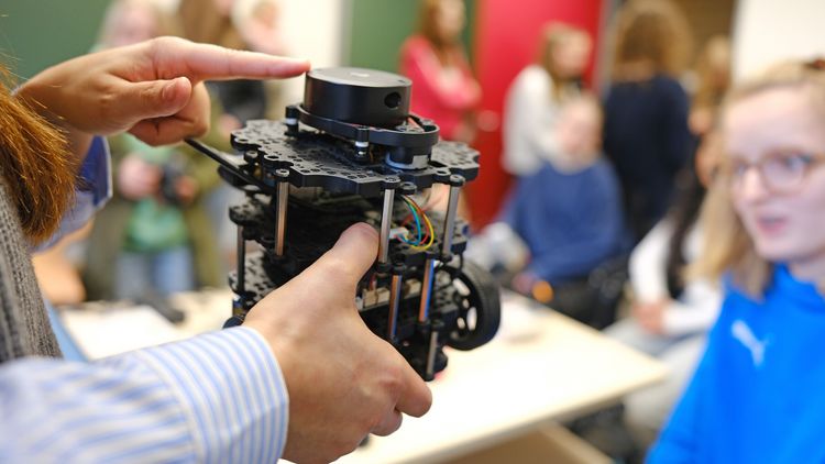 Eine Person hält eine kleine Apparatur, womöglich ein Roboter, in ihren Händen und zeigt sie einer Gruppe von Kindern.
