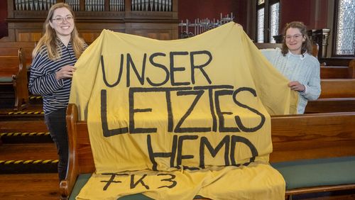 Das Bild zeigt Wencke Bammann und Mafalda Nogueira. Sie halten ein überdimensioniertes gelbes T-Shirt mit der Aufschrift "Unser letztes Hemd. Fakultät 3" hoch. Im Hintergrund sind eine Orgel und Sitzbänke des alten Lehrerseminars zu erkennen.