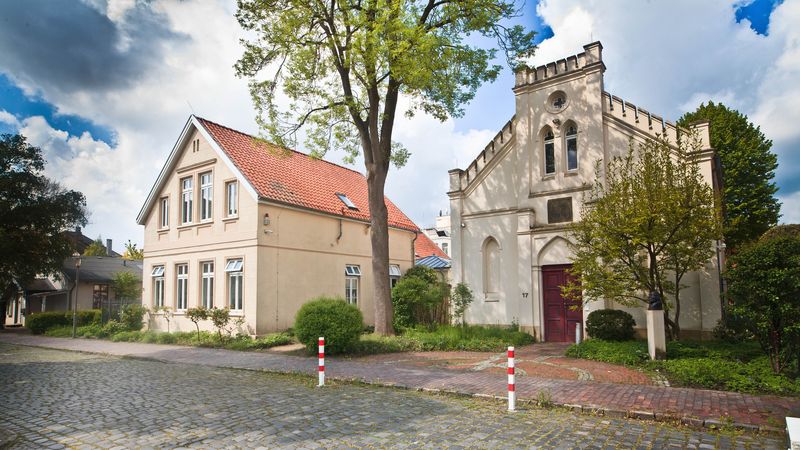 Zwei Gebäude im Gründerzeitstil an einer Kopfsteinpflasterstraße, umgeben von Bäumen und im Hintergrund bewölkter Himmel. 