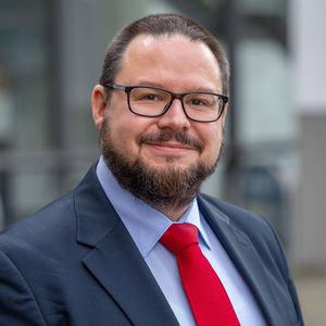 Portraitfoto von Professor Dr Torsten Jantsch, der eine schwarze rechteckige Brille, ein dunkelblaues Jacket mit hellblauem Hemd und eine rote Krawatte trägt. Er lächelt in die Kamera, hat kurze dunkle Haare und einen kurzen dunklen Bart, der Wangen, Kinn und Oberlippe bedeckt.
